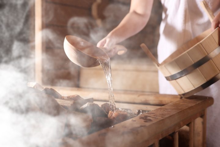 Woman pouring water onto stones surrounded by steam in sauna, closeup.