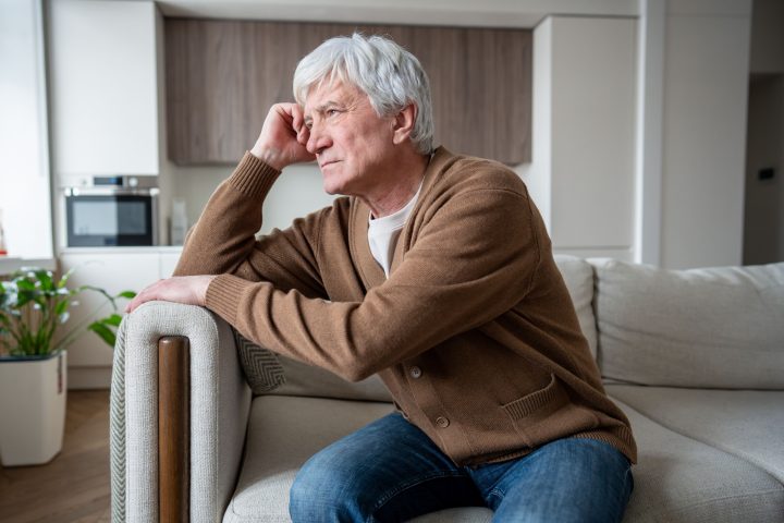 Aging senior man sits alone in empty living room staring vacantly, struggling with memory loss. Alzheimer confusion, chronic loneliness, cognitive decline, abandoned elder, neurological deterioration